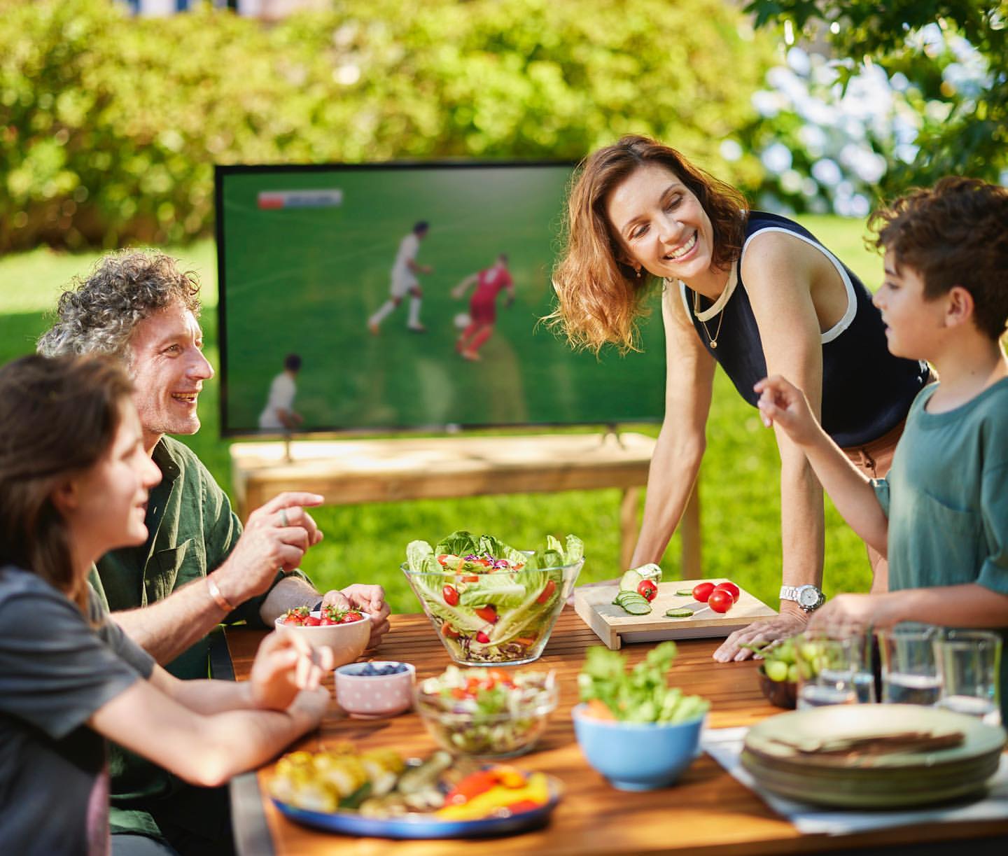 Family of four watching sports on TV in the backyard while eating salad and sliced vegetables on a sunny day outdoors.