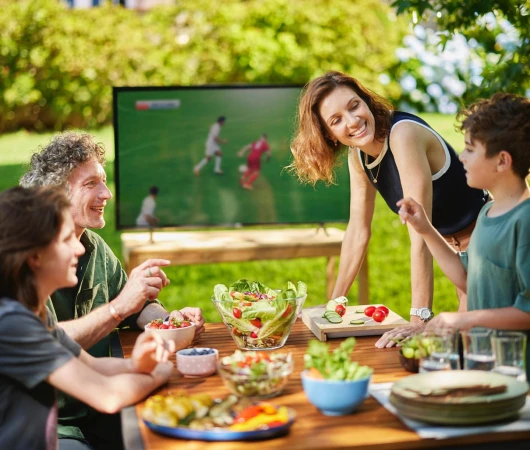 Family of four watching sports on TV in the backyard while eating salad and sliced vegetables on a sunny day outdoors.
