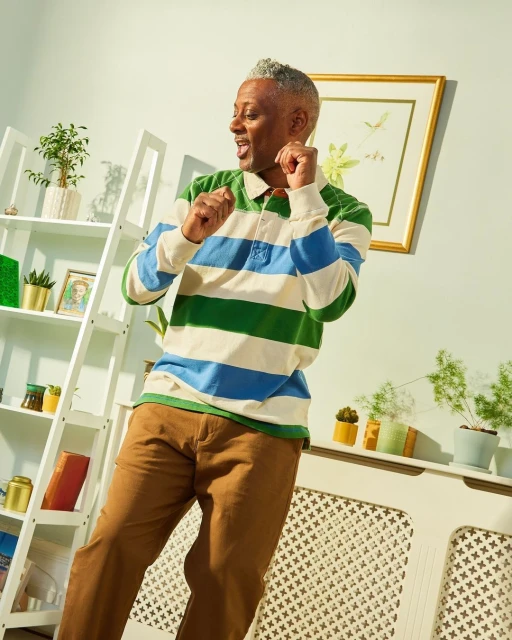 An adult male with grey hair in a blue and green striped shirt poses in an indoor setting next to shelves with various objects and plants.