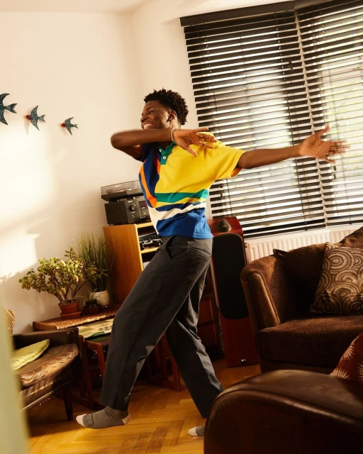 A young Black man indoors, dancing with arms outstretched, wearing a colorful shirt and grey trousers, smiling, in a cozy living room setting.