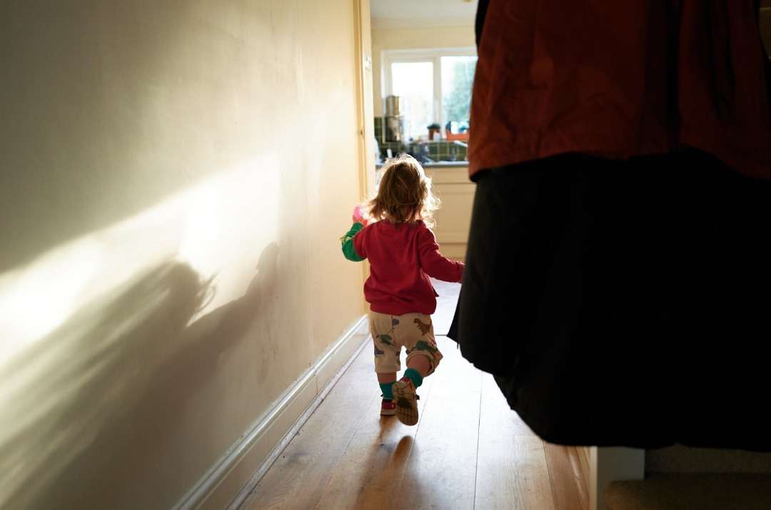 Blond toddler walks down a sunlit hallway wearing a pink sweater, patterned pants, and brown shoes, seen from behind and partially obscured.