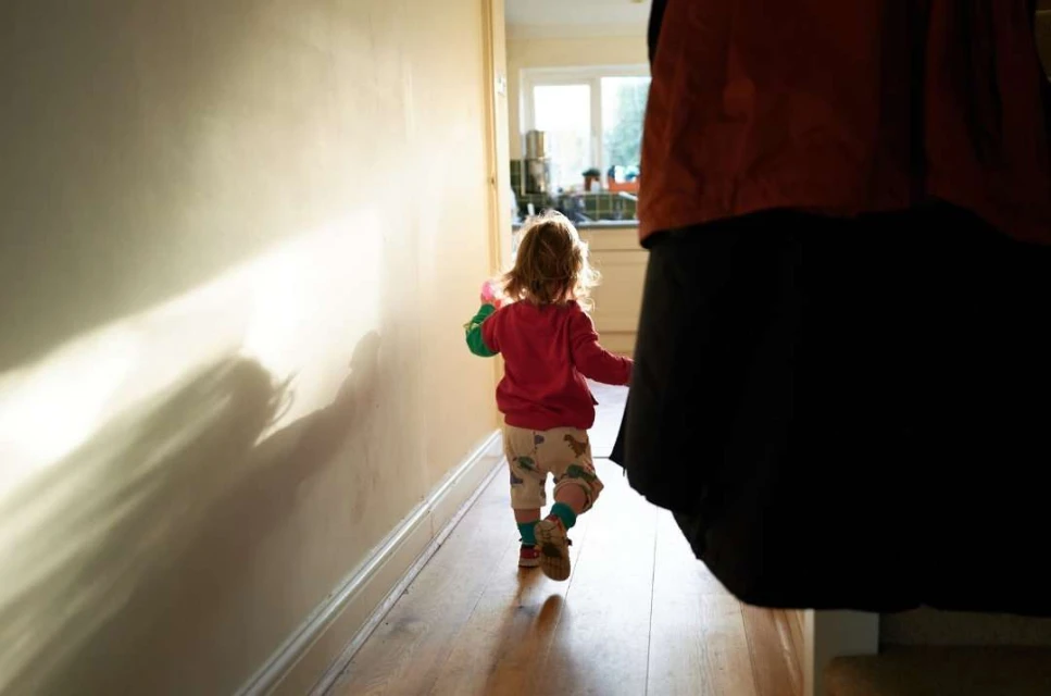 Blond toddler walks down a sunlit hallway wearing a pink sweater, patterned pants, and brown shoes, seen from behind and partially obscured.