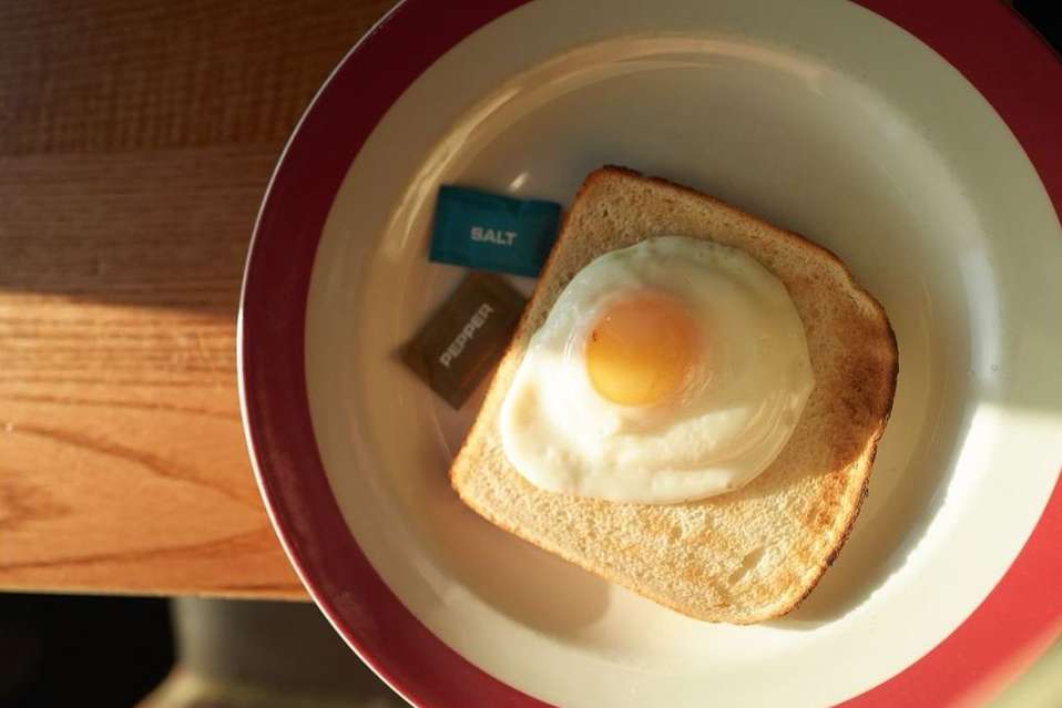 Overhead shot of fried egg on toast with salt and pepper packets on a red-rimmed plate, bright sunlight, and neutral tabletop.