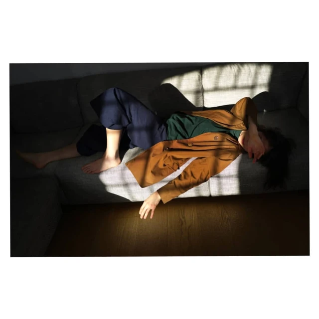 A woman in a jacket and trousers reclines on a couch in the sun. The shadows from the sun create a pattern on the furniture and floor.