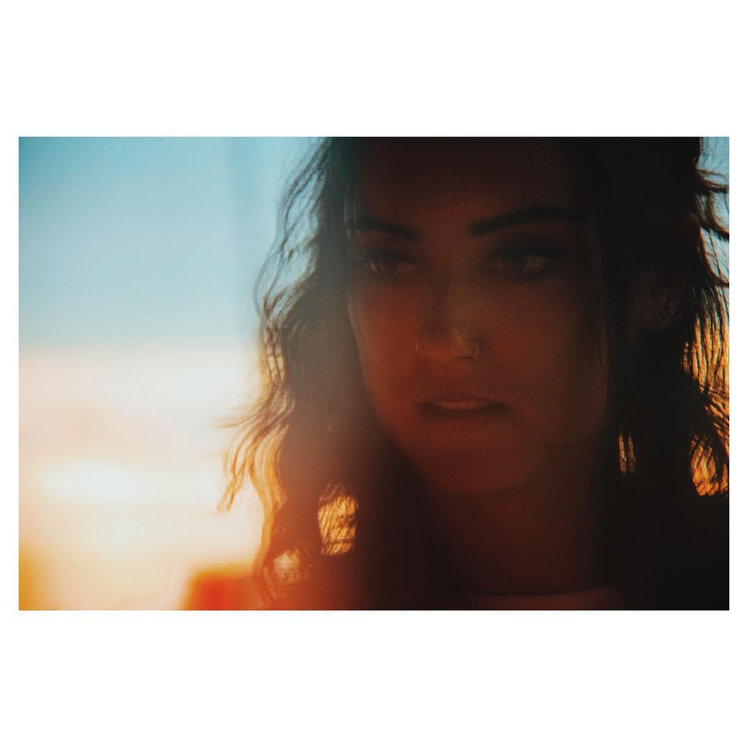 Close-up of a dark-haired young adult female with a nose ring looking off-camera with a blue and orange sunset background.