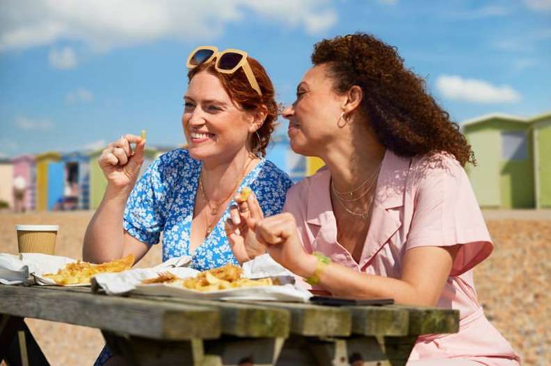Two women laugh together at an outdoor picnic table laden with food. Beach huts and blue sky visible in the bright background.