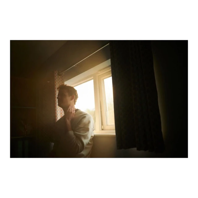 Adult male in a light sweater stands near a window with drawn curtains, bathed in soft, natural light, creating a contemplative mood.