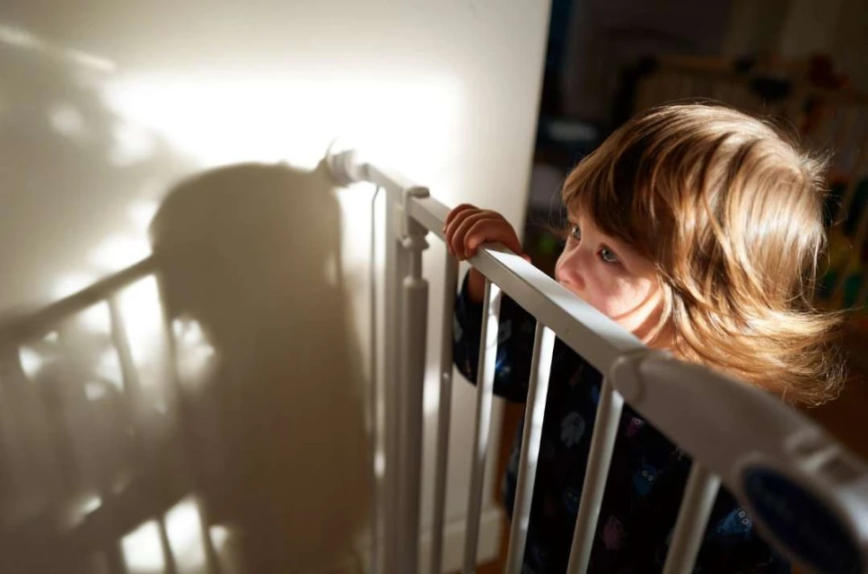 Young child with long brown hair peers over a white safety gate in a brightly lit room, casting a large shadow on the wall.
