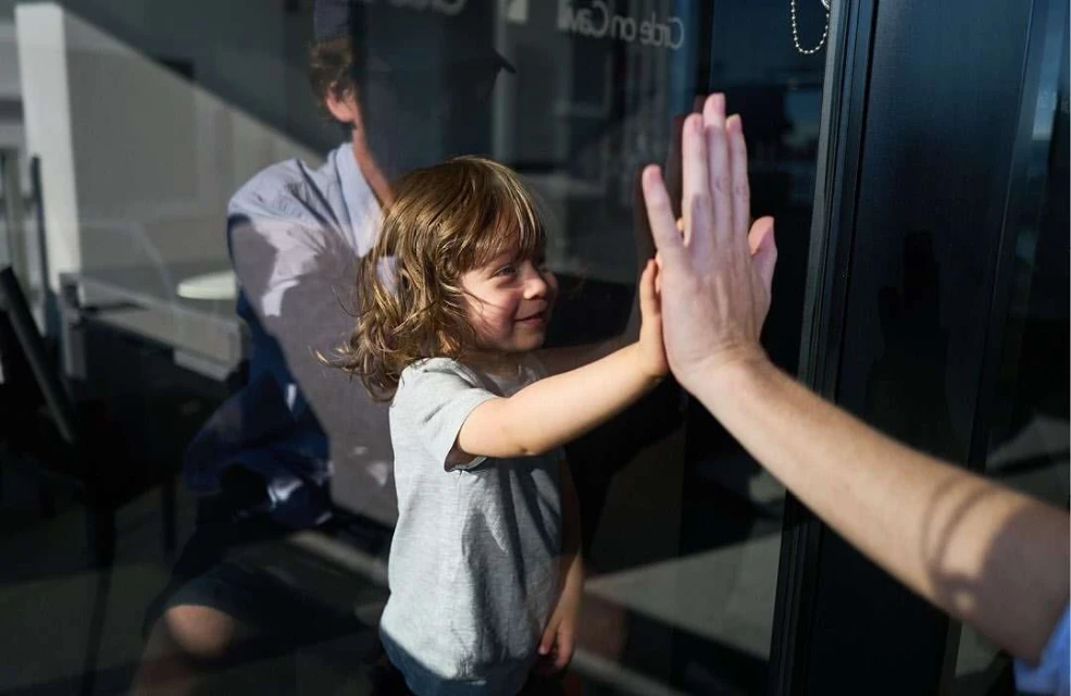 A young child touches hands through glass with an adult; a man stands in the background in an indoor setting with natural lighting.