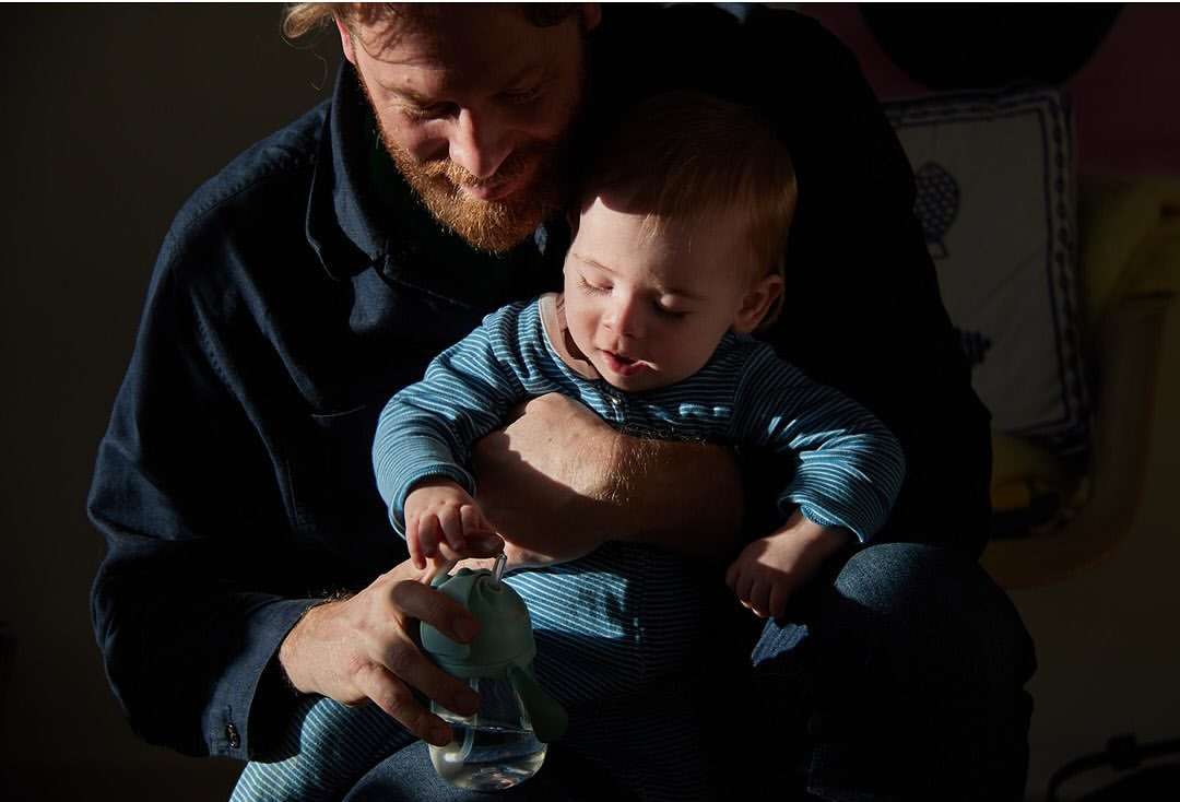 A father and baby sit indoors in dappled sunlight. The man holds a baby bottle as his child sits on his lap in a cozy domestic scene.