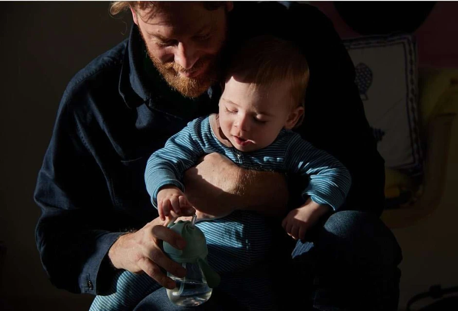 A father and baby sit indoors in dappled sunlight. The man holds a baby bottle as his child sits on his lap in a cozy domestic scene.