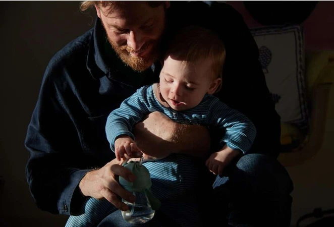 A father and baby sit indoors in dappled sunlight. The man holds a baby bottle as his child sits on his lap in a cozy domestic scene.