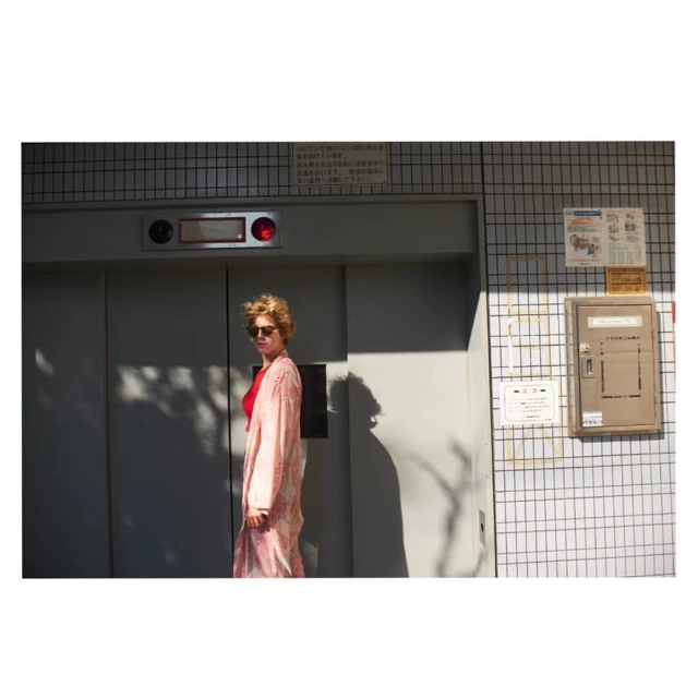Adult female standing in front of a closed elevator in a pink patterned robe, sunglasses, and red top in an outdoor urban setting.