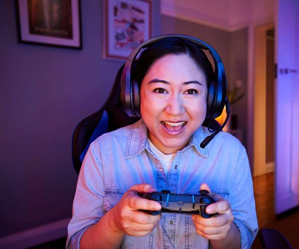 Enthusiastic Asian woman gamer with headset smiles, holds controller. Indoor setting, blue and red lighting, gaming chair visible.