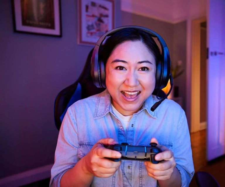 Enthusiastic Asian woman gamer with headset smiles, holds controller. Indoor setting, blue and red lighting, gaming chair visible.
