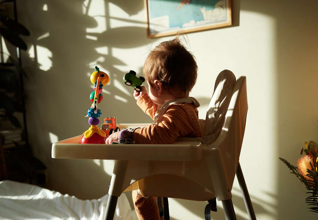 A baby sits in a high chair in a sunlit room, playing with a toy car and a colorful stacked toy. Indoor scene, natural light, calm mood.