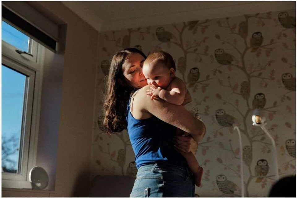 A woman in a blue tank top and jeans holds a baby in a diaper indoors, with a window to the left and owl-themed wallpaper behind them.