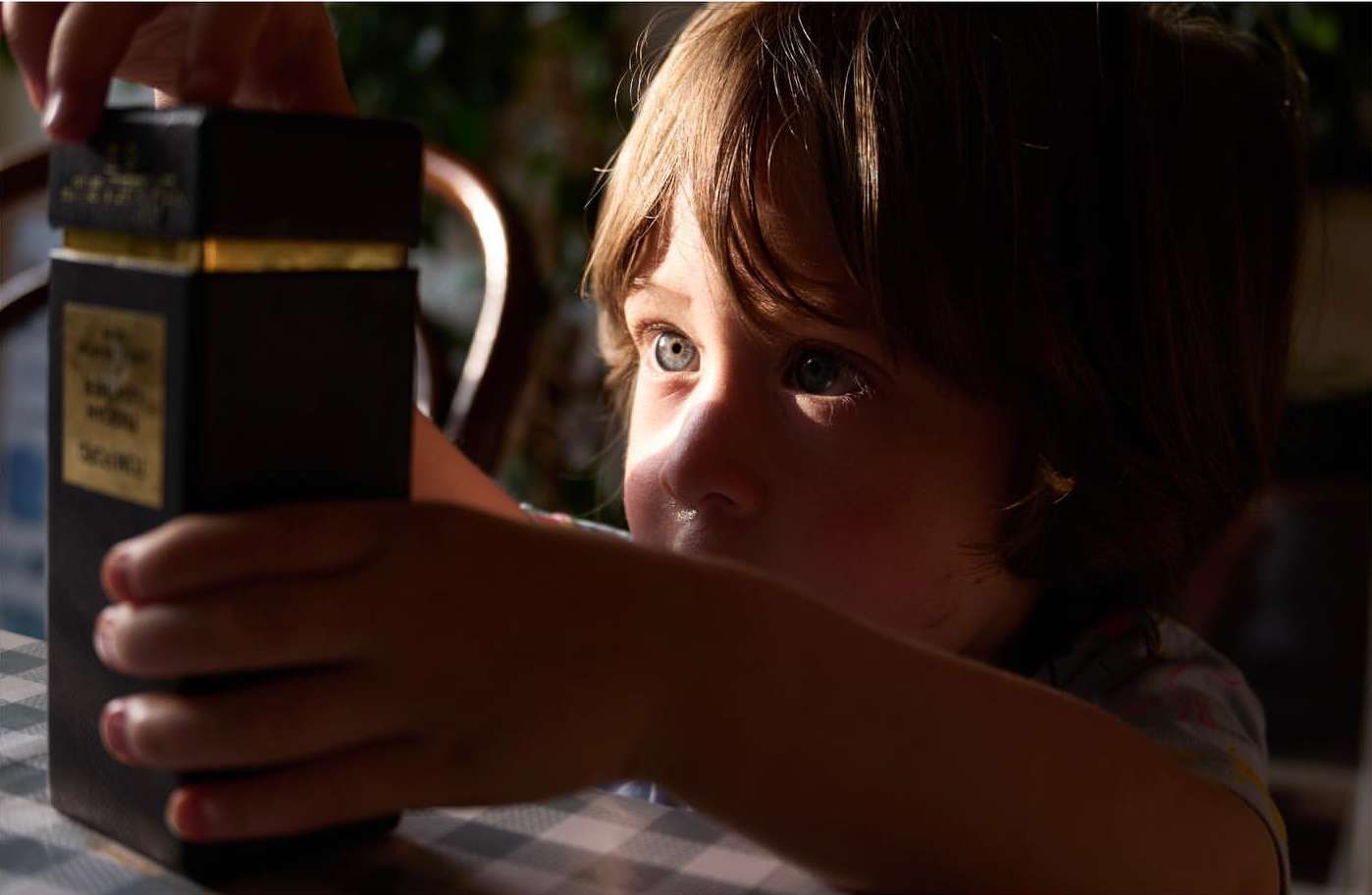 Close-up of a child with blue eyes holding a dark rectangular box with gold accents inside a home environment, looking towards the light.