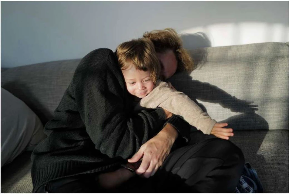 Mother and child share a warm embrace on a grey couch, with sunlight streaming through a window casting shadows on the wall.