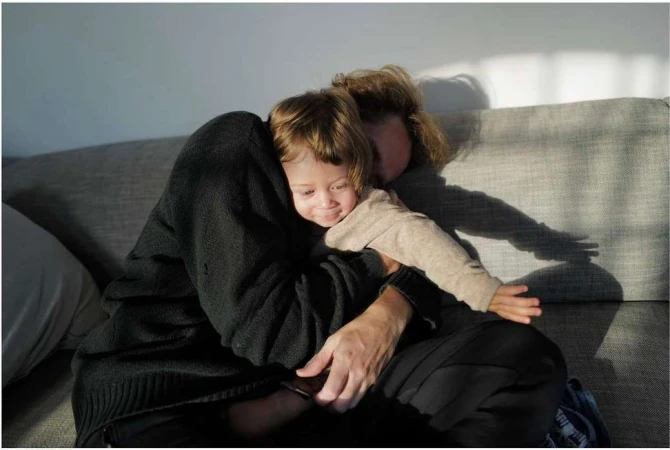 Mother and child share a warm embrace on a grey couch, with sunlight streaming through a window casting shadows on the wall.
