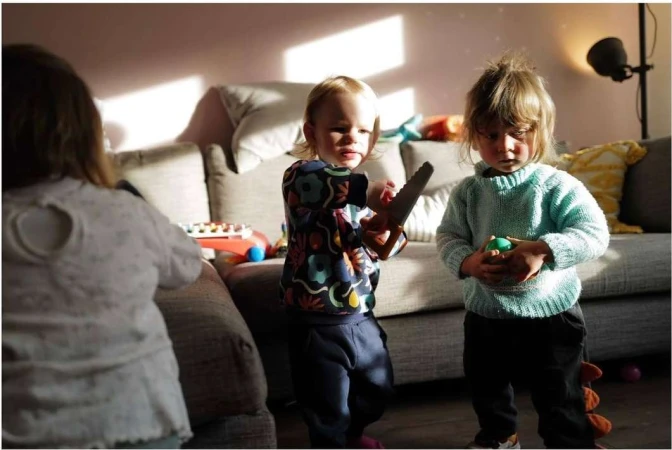 Two young children indoors playing with toys and interacting on a couch, bathed in soft, natural light from a nearby window.