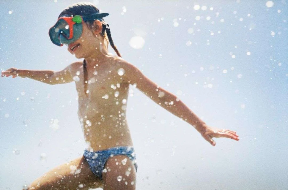 A young child in a blue patterned swimsuit and swimming goggles is shown outdoors playing in the water on a sunny day.