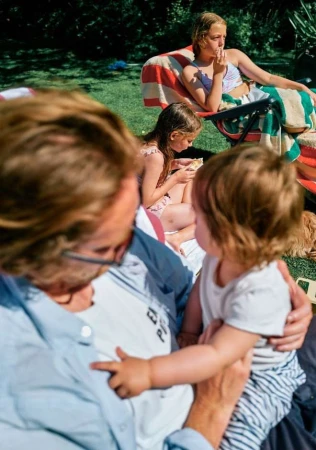 A family relaxes outdoors on a sunny day. A toddler interacts with an adult while two girls lounge on the grass in the background.