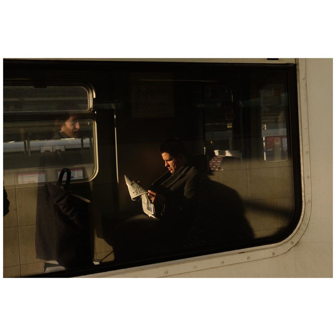 Person reading a newspaper on a train, captured through the window from outside. Sunlight and shadow create visual contrast.