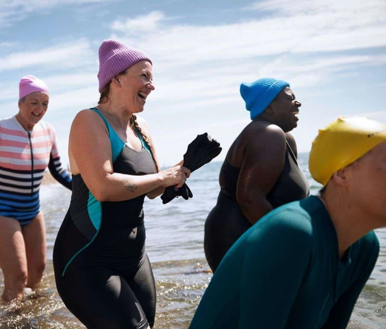 Group of women in swimsuits and swimming caps standing in shallow ocean water on a sunny day, laughing and preparing to swim.