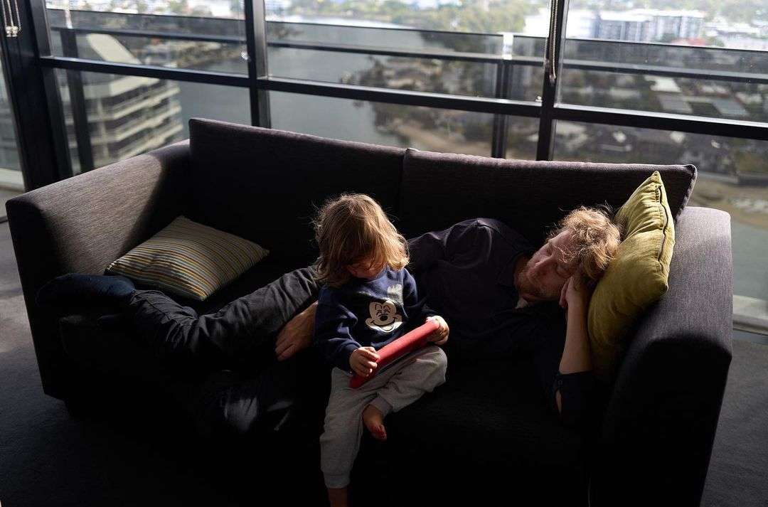 A young child sits on a dark grey sofa in a modern apartment, using a tablet while an adult sleeps next to them near a large window.