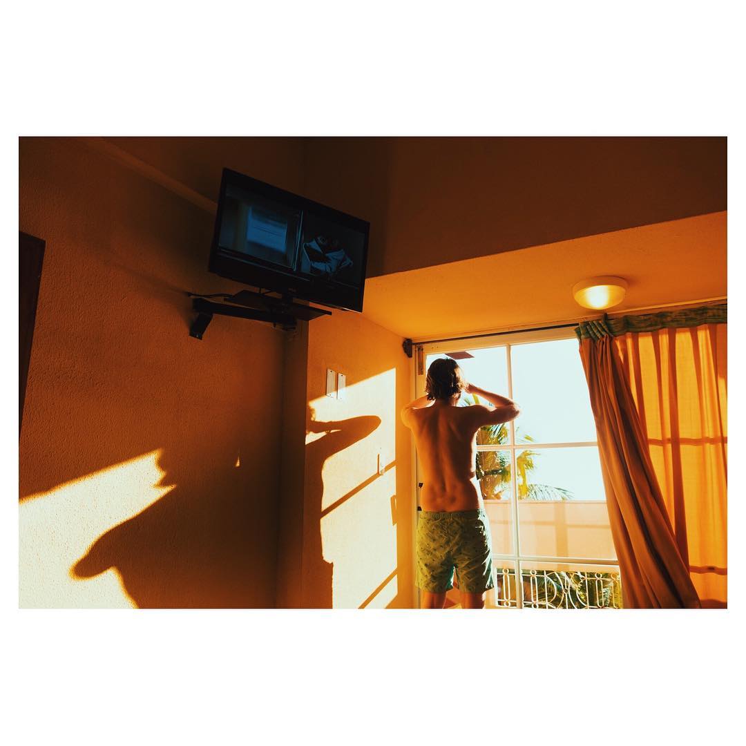 A man stands shirtless in shorts in a room with an open door to a balcony, looking out into bright sunlight, with a television on the wall.