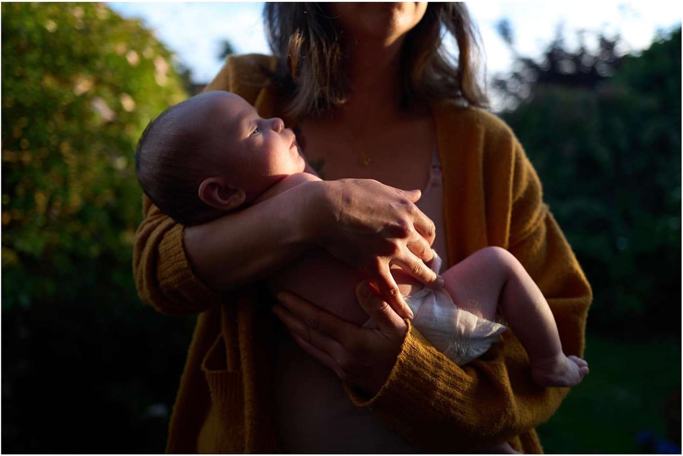 A person holds a baby in their arms, outdoors, with soft, diffused lighting highlighting the baby's face. The background is blurry foliage.