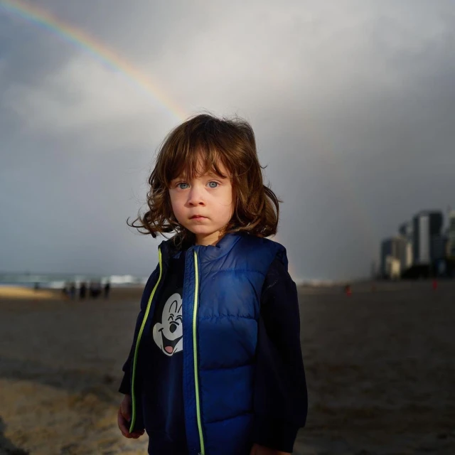 Young child stands on a beach near the ocean under a colorful rainbow in the sky in an eye-level, medium shot with soft, diffused light.