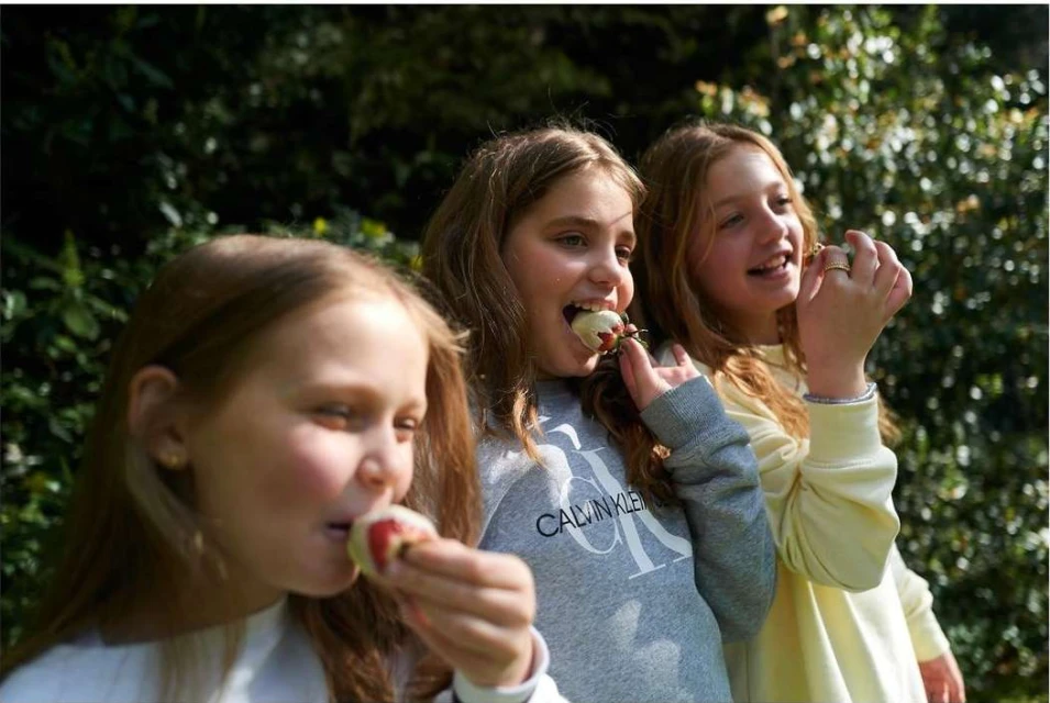 Three young girls eating fruit outdoors in a sun-dappled setting, wearing casual clothing, capturing a moment of youthful enjoyment.