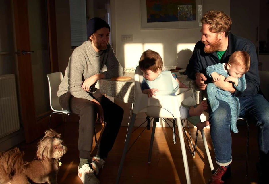 Two adult males and two infants are gathered at a table in a sunlit room, accompanied by a small dog at their feet. 