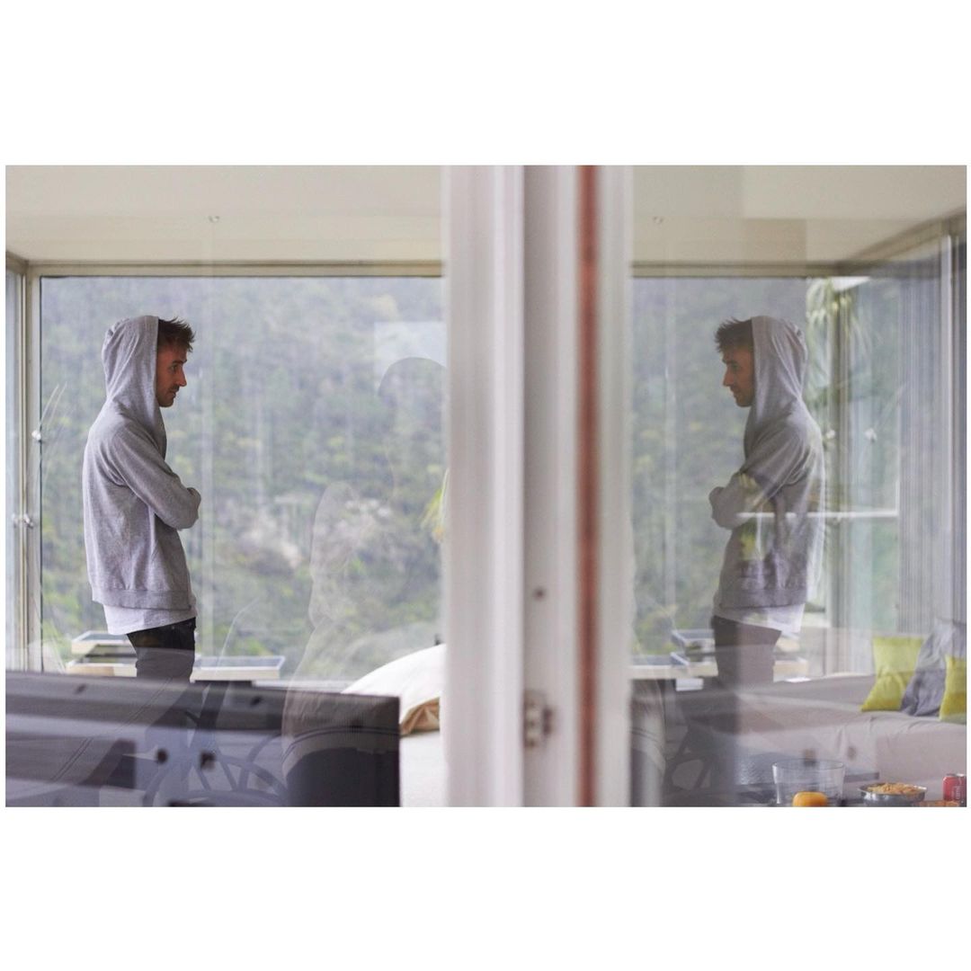 An adult male stands indoors near a large glass window, looking off at a mountainous view. The man is wearing a grey hooded sweatshirt.