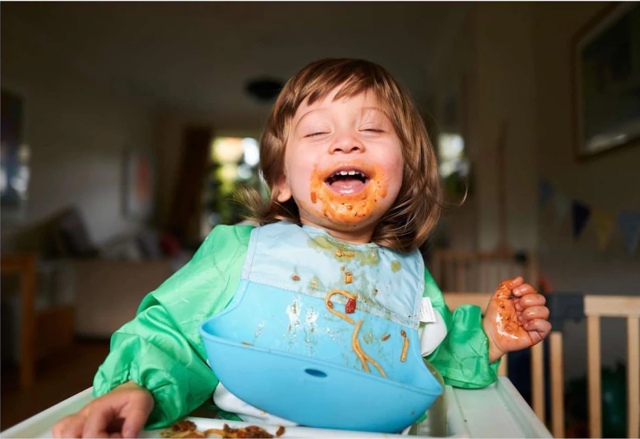Child with messy face covered in spaghetti sauce, wearing a blue bib and green sleeves, sitting in a high chair in a bright indoor setting.