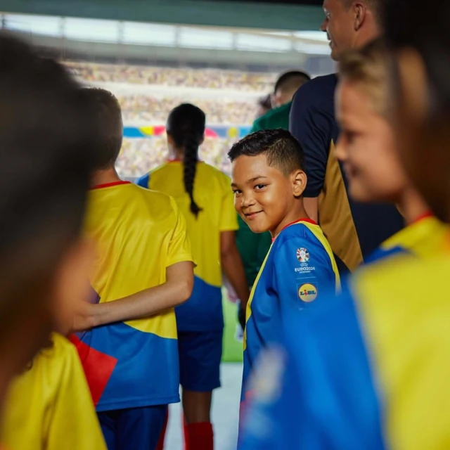 Smiling boy in blue soccer jersey surrounded by kids with stadium backdrop. Youth sports, teamwork, and fan spirit are visible.