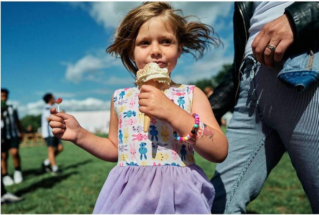 A young child eating an ice cream cone outdoors, wearing a patterned dress with a purple skirt and holding a small lollipop.