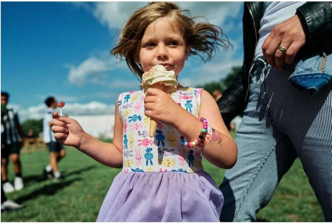 A young child eating an ice cream cone outdoors, wearing a patterned dress with a purple skirt and holding a small lollipop.