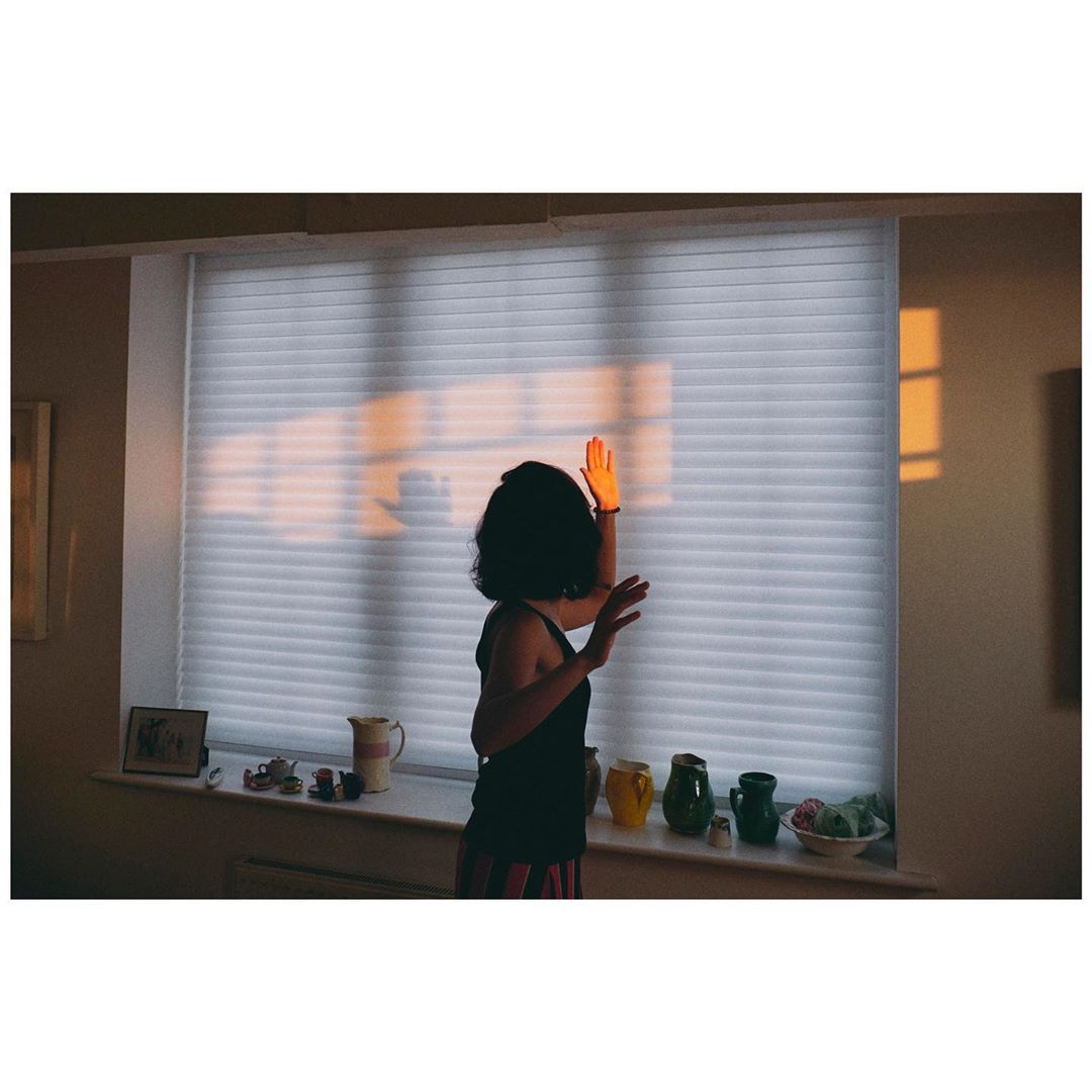 Backlit woman raises hand in a sunlit room with white window blinds, decorative kitchenware on a windowsill, and warm, golden light.