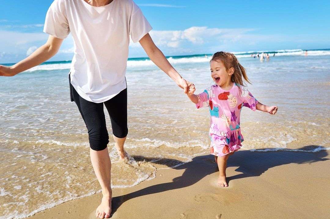 Happy little girl running on the beach with her mom on a sunny day. The ocean waves are crashing in the background under a clear sky.
