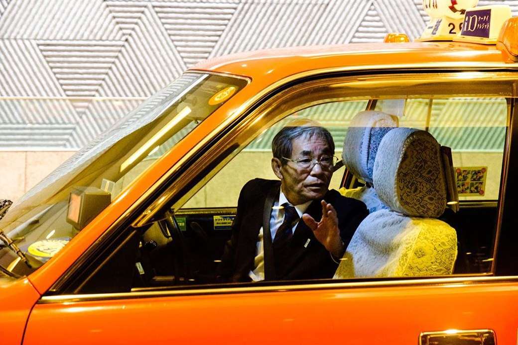 Elderly Asian taxi driver in business suit with glasses sitting inside an orange taxi in city. Geometric patterned wall background.