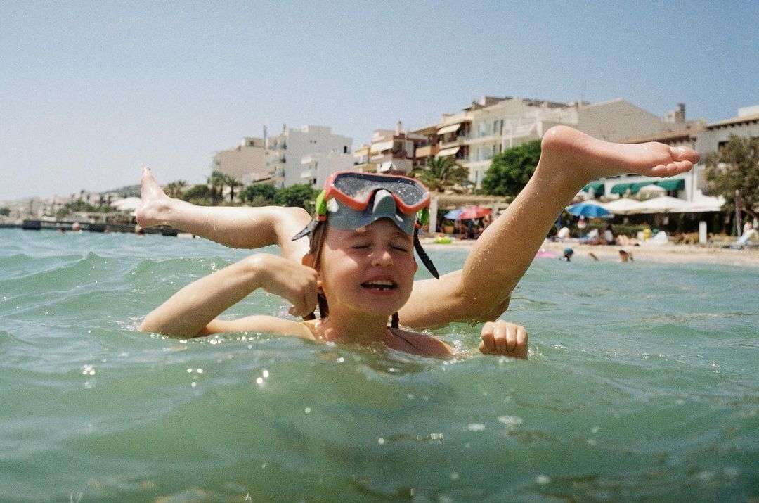 Child with snorkel mask in ocean water, sunny day at beach with buildings and palm trees in background, fun summer vacation activity.