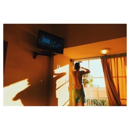 Shirtless man wearing swim trunks standing by a bright window in a hotel room with warm sunlight