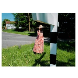 Woman in a red and white striped dress and wide-brimmed hat standing by a rural roadside