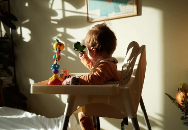 Young child seated in a high chair playing with toys under warm sunlight by Kell Mitchell in London