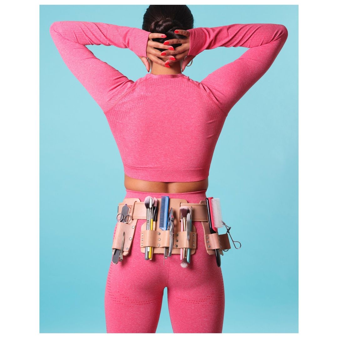 Back view of a female makeup artist wearing a pink athletic outfit and a makeup tool belt with various cosmetic brushes and hair tools.