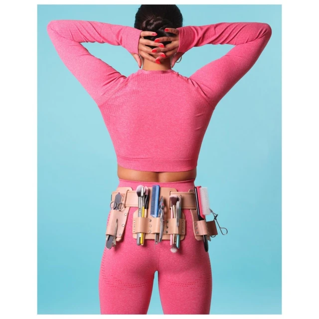 Back view of a female makeup artist wearing a pink athletic outfit and a makeup tool belt with various cosmetic brushes and hair tools.
