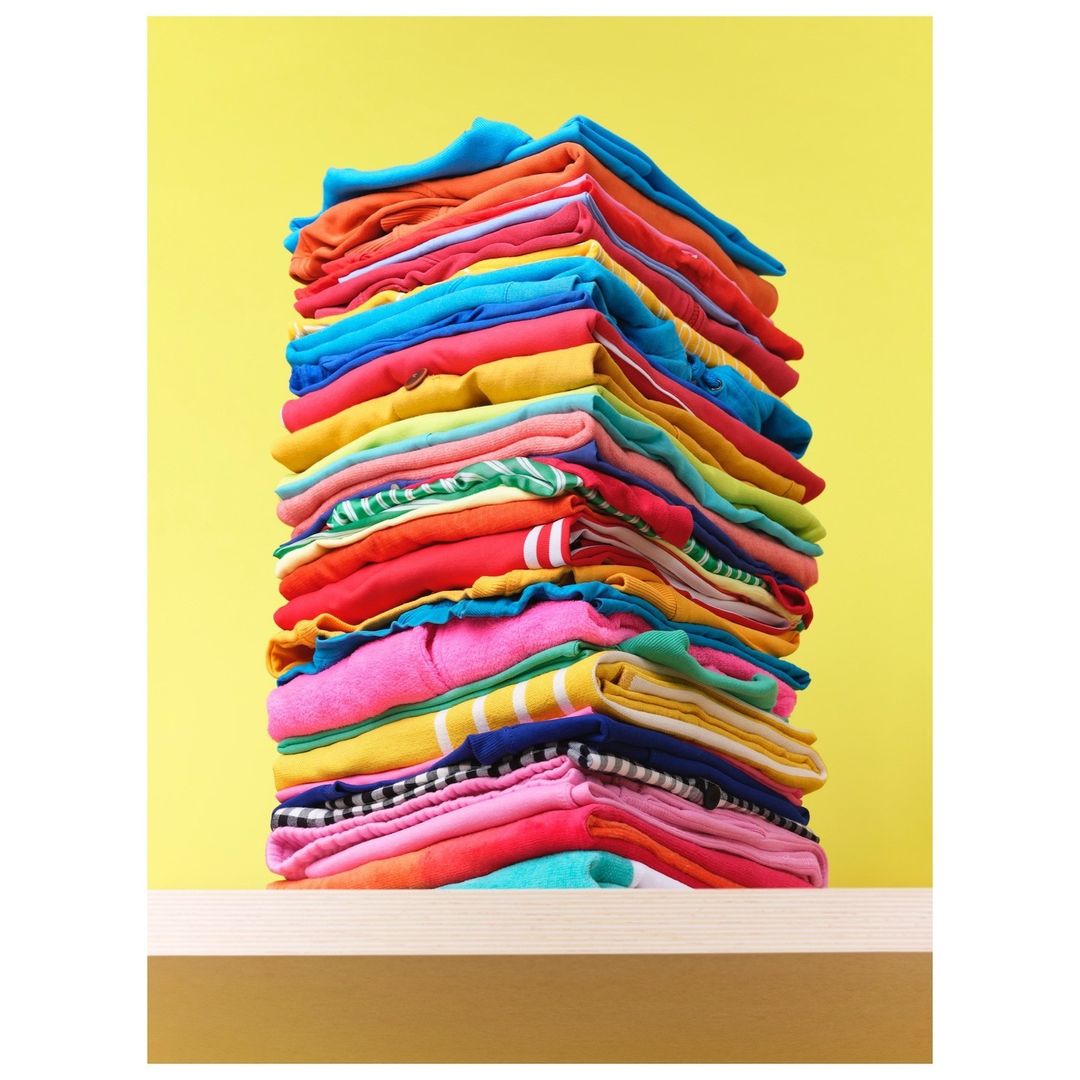 Colorful stack of folded clothes on a light wooden shelf against a vibrant yellow background, creating a cheerful and organized visual.